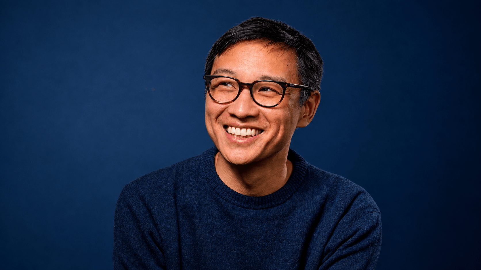 Homem sorrindo, de óculos e suéter azul, diante de um fundo marinho.